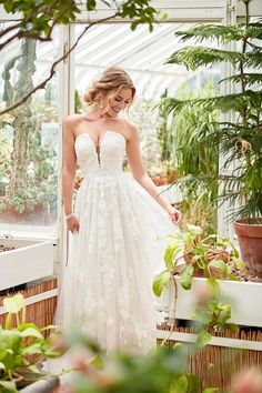 a woman in a white wedding dress standing next to a greenhouse filled with potted plants