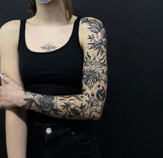 a woman with tattoos on her arm wearing a face mask and black tank top standing in front of a dark background