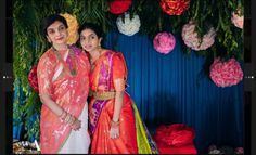 two women standing next to each other in front of flowers and greenery on the wall