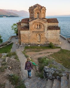 a woman walking up some steps towards an old church by the water with mountains in the background