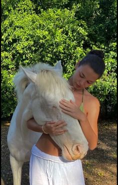 a woman is hugging a white horse in front of some green bushes and shrubbery