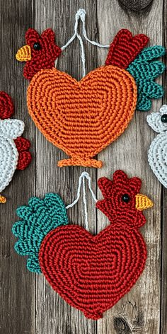 three crocheted roosters hanging from strings on a wooden surface with yarn in the shape of a heart