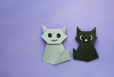 two origami cats sitting next to each other on a purple surface with one cat looking at the camera