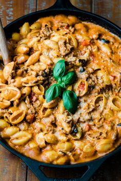 a skillet filled with pasta and sauce on top of a wooden table next to a spoon