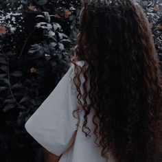 a woman with long curly hair standing in front of some bushes and flowers looking at the sky