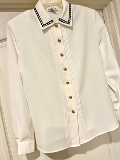 This is a beautiful classic 1990's soft-white wrinkle polyester perfectly tailored blouse with a row of 4 tucks on each side of the opening.  Beautiful metal gold buttons down the front and braided embroidery navy trip to the collar.  Travel in style with no-wrinkle long sleeves.  Chest measures 22 inches across at armpits and length 26 inches. Several marks to left of collar...see last photo.  This is new with tags and extra metal gold button. Mailed priority. Braided Embroidery, Tailored Blouse, Embroidery Collar, Navy Embroidery, Free Front, Collar Blouse, Gold Buttons, Wrinkle Free, Soft White