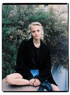 a woman sitting on top of a cement wall next to trees and bushes with her legs crossed