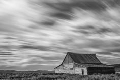 a black and white photo of an old barn in the middle of nowhere with clouds overhead