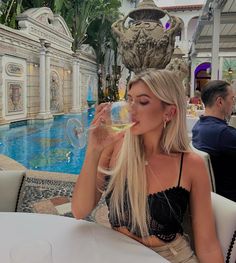 a woman sitting at a table drinking from a wine glass in front of a pool