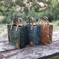 Three small waxed canvas totes on a picnic table in a park. Handcrafted byRoad Dirt and Pie and available in green, gray, and brown colors. Outdoor Canvas Tote Bag With Reinforced Handles, Eco-friendly Waxed Canvas Bags For Daily Use, Waxed Canvas Bag For Daily Use, Everyday Waxed Canvas Bag With Reinforced Handles, Eco-friendly Waxed Canvas Bag With Canvas Lining, Everyday Duck Canvas Bag With Waxed Finish, Waxed Coated Canvas Tote Bag, Everyday Duck Canvas Bags With Waxed Finish, High-end Rectangular Canvas Bag With Waxed Finish