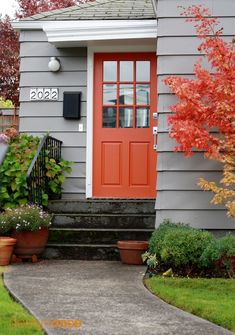 a red door is on the side of a gray house with potted plants and trees