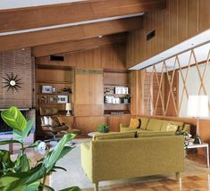 a living room with wood paneling and green furniture in the center, surrounded by large potted plants