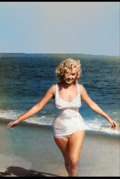 a woman in a white bathing suit walking on the beach with her arms spread out