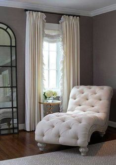 a white chaise lounge chair sitting in front of a window with drapes on the windowsill