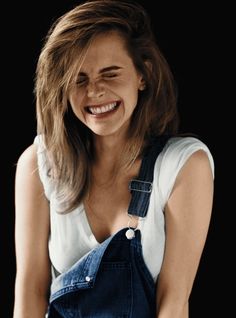 a smiling woman in overalls with her hands on her hips and one eye open