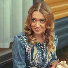 a woman with blonde hair and blue shirt holding a cell phone in her hand while sitting down