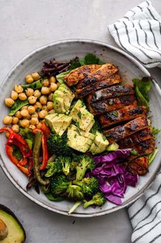 a white plate topped with meat, vegetables and chickpeas next to avocado