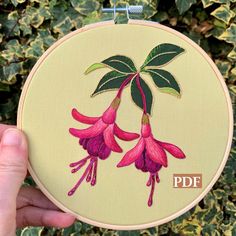 a hand holding up a embroidery hoop with pink flowers on it and green leaves in the background