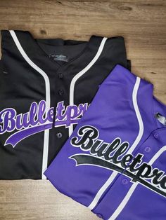 two purple and black baseball jerseys laying on top of a wooden floor next to each other