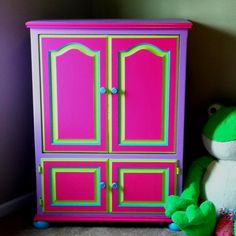 a green stuffed frog sitting next to a pink and green armoire with two doors