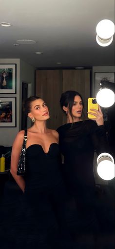 two women are taking a selfie in the mirror with their cell phone and one is wearing a black dress