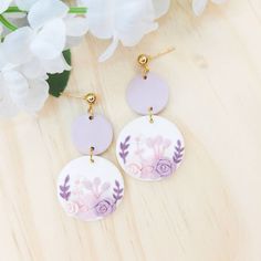 two purple and white earrings with flowers on them sitting next to a flower bouquet in front of a wooden surface