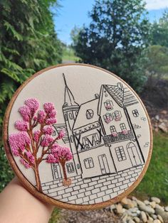 a hand holding up a small embroidery art piece with pink flowers on it and a castle in the background