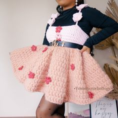 a woman wearing a pink skirt and black top with red flowers on it, standing in front of a white wall