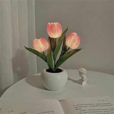 a white vase filled with pink flowers sitting on top of a table next to an open book