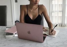 a woman sitting at a table with an apple laptop in front of her, writing on a notepad