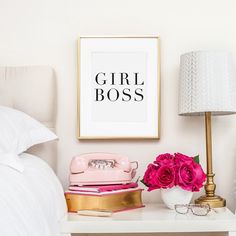 a white bed topped with pink flowers next to a lamp and a framed print on the wall