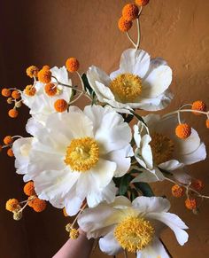 white and yellow flowers are in a vase on a brown tablecloth with a person's hand holding it