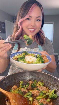 a woman holding a bowl of broccoli and chicken with chopsticks in her hand