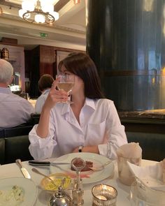 a woman sitting at a table holding a wine glass in front of her face with food on it