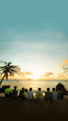 group of people sitting on the beach watching the sun go down over the ocean with palm trees