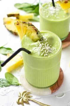 two glasses filled with green smoothie next to sliced pineapples and other fruit