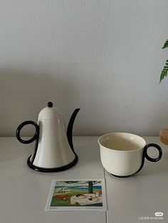 a white coffee pot sitting next to a cup on top of a table near a plant