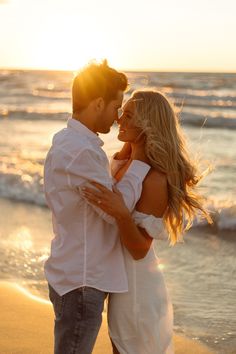 a man and woman are standing on the beach at sunset with their arms around each other