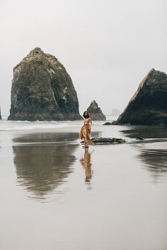 a woman standing on top of a sandy beach next to two large rocks in the ocean