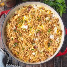 a large skillet filled with pasta and meat on top of a wooden table next to parsley