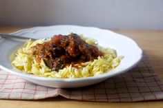 a white bowl filled with pasta and meat on top of a wooden table next to a red checkered napkin