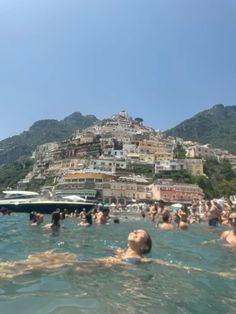 many people are swimming in the water near a hill with houses on it and boats