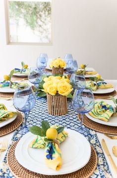 the table is set with yellow flowers and blue glassware, gold place settings, and wicker napkins