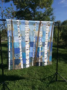 a quilt hanging on a stand in the grass with trees behind it and blue sky above