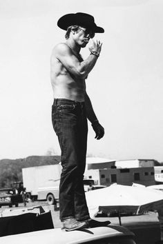 a shirtless man standing on the hood of a car while talking on a cell phone