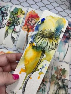 a person holding up some watercolor paper with flowers on it and another piece of artwork in the background