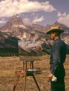 a man standing in front of an easel holding a paintbrush and looking at the mountains