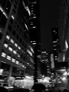 black and white photograph of city buildings at night with cars passing in the foreground