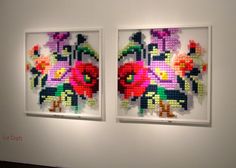 two cross stitch paintings on display in a white walled room with red and yellow flowers