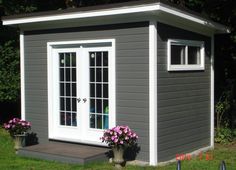 a small gray shed with white doors and windows on the side, next to some potted flowers